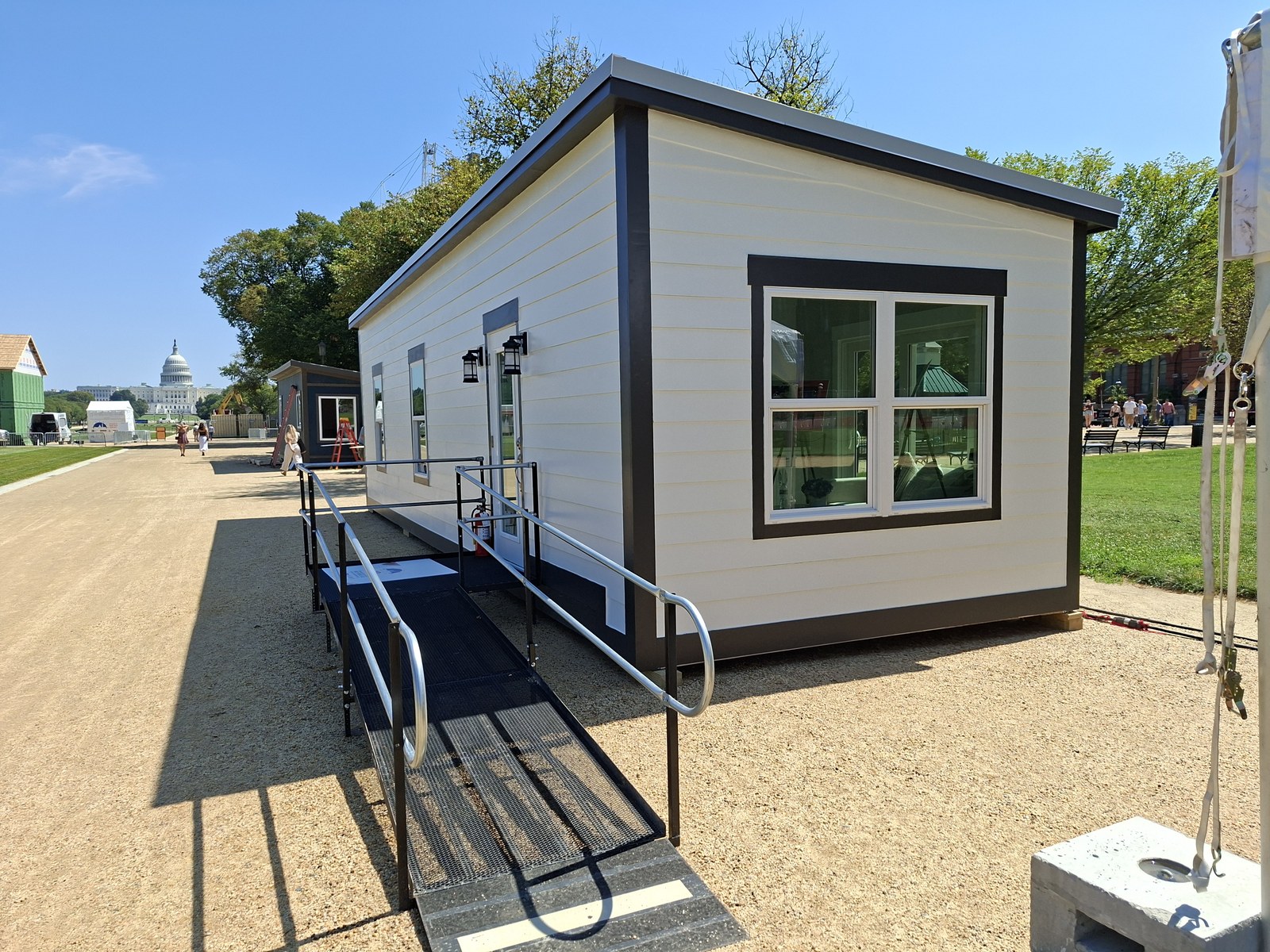Conventional modular dwelling exhibited at the 2025 HUD Innovative Housing Showcase on the National Mall in Washington DC. White siding with dark trim, traditional manufactured-home aesthetic, accessibility ramp at front. US Capitol dome visible in the distance at left.