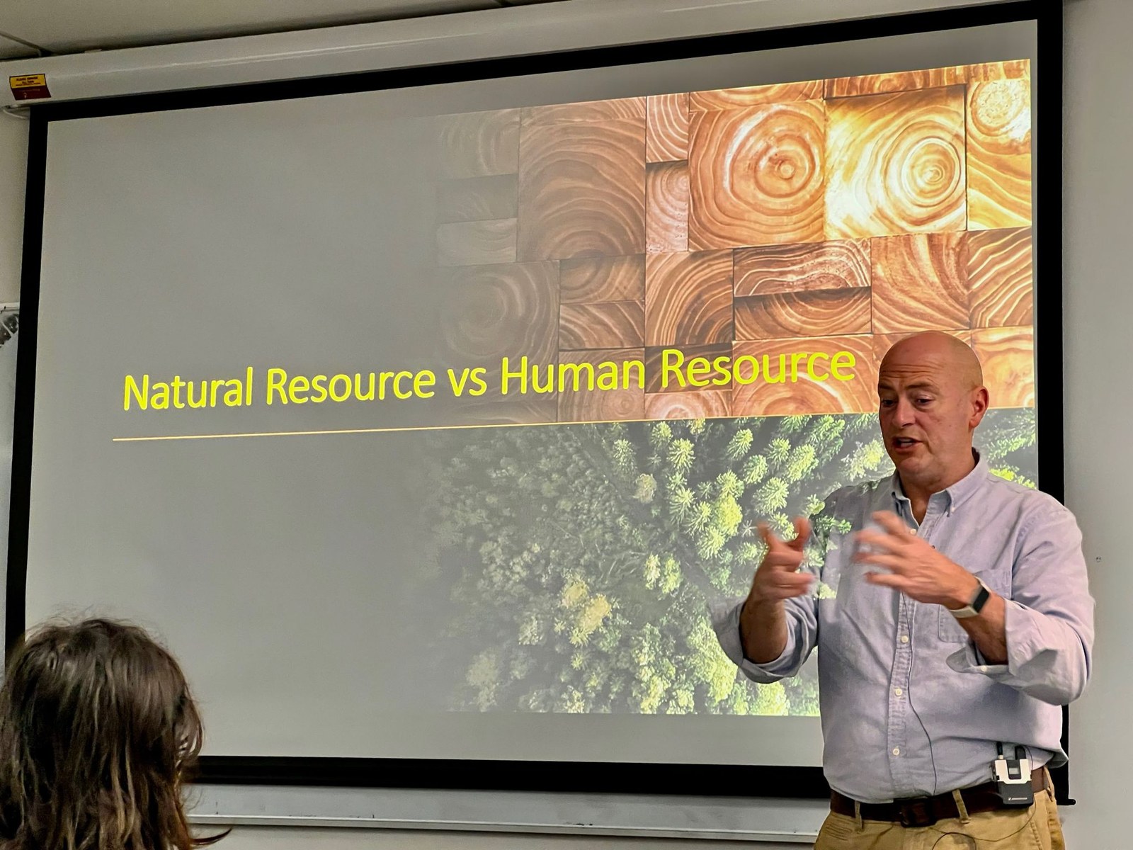 The lecture slide titled Natural Resource vs Human Resource visible on the projection screen behind Murphy O'Neal, who is mid-explanation. The slide contrasts cut timber end-grains showing the growth rings of mature trees with an aerial view of forest canopy below.