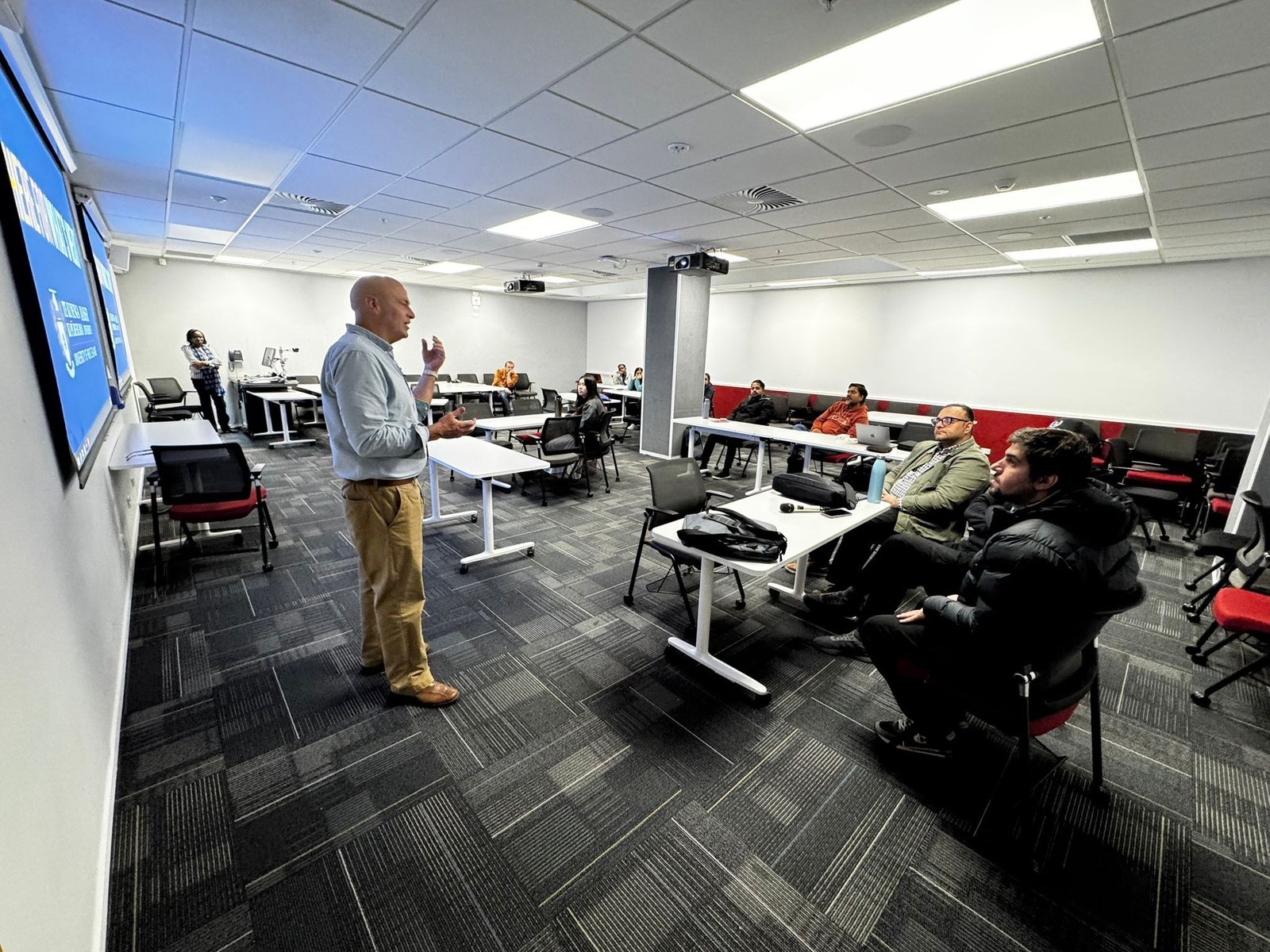 Murphy O'Neal presenting at the Massey University School of Built Environment Research Seminar, August 5 2025. Wireless mic transmitter visible on belt. Multiple attendees seated at tables with laptops open. Massey University HERE FOR WHAT'S NEXT branding visible on lecture room screen.