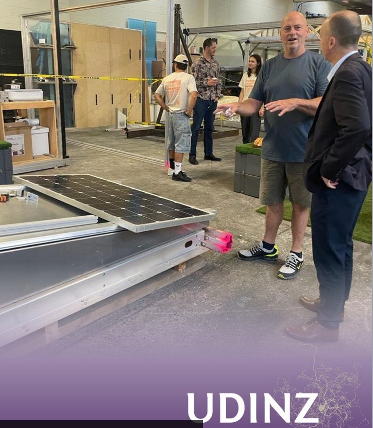 Murphy O'Neal explaining the solar roof integration to a UDINZ attendee at right. Solar panels visible mounted on aluminum extrusion roof framework in the foreground.