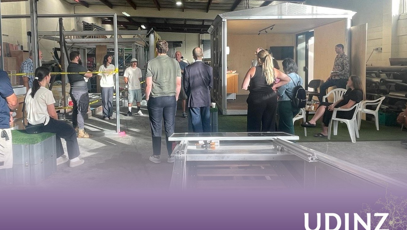 UDINZ Summer Site Tour at the Adaptable Structures facility, 9 Fremlin Place Avondale Auckland, 18 February 2025. Approximately twelve UDINZ urban-development professionals visible in the warehouse touring the Anyplace facility. An Anyplace module visible at right with sliding door open. A solar roof component on the floor in foreground.